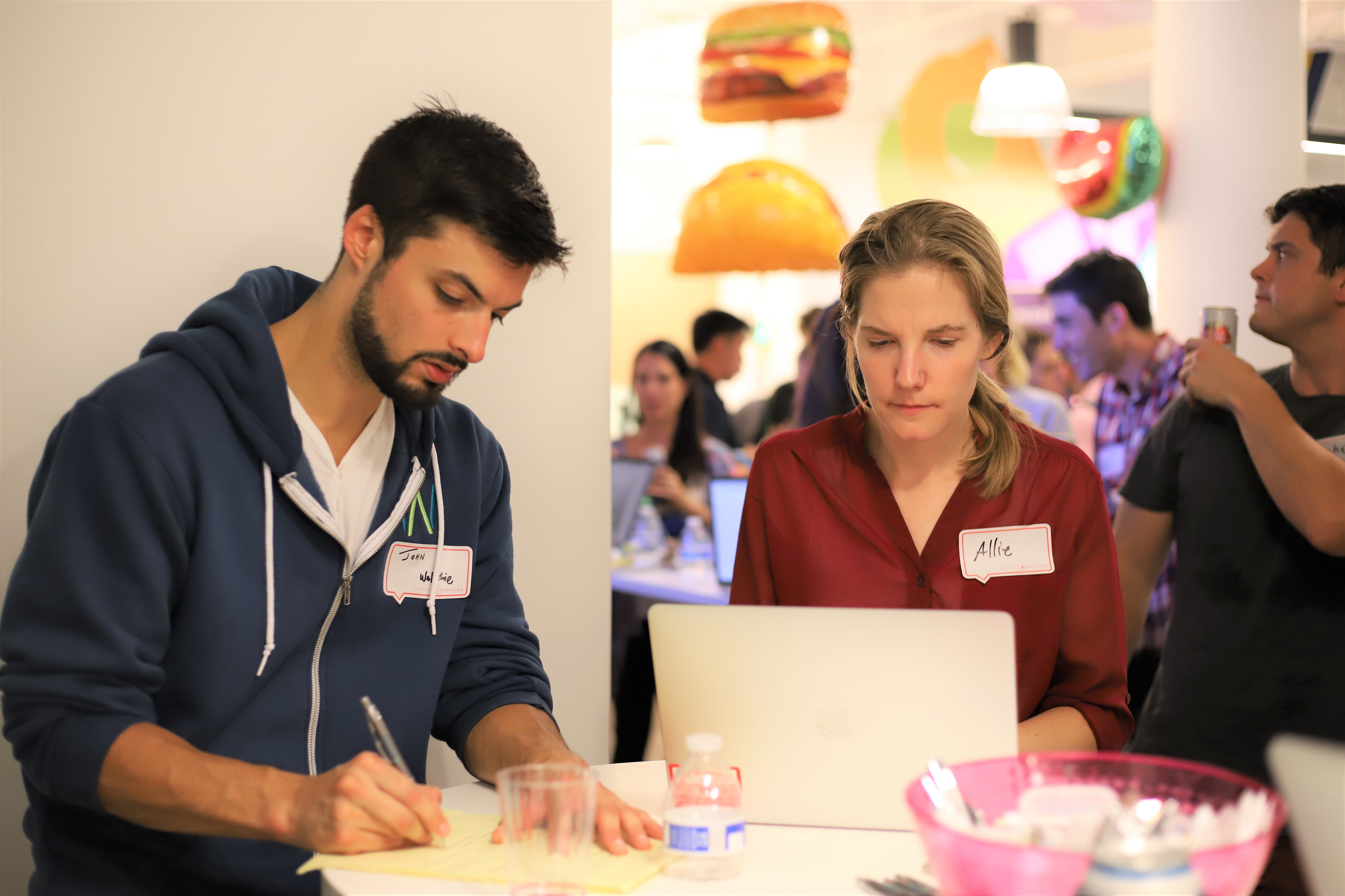 This is a picture of two users testing a product at a user testing event. This is a photo of a man and a woman wearing nametags and writing feedback about a software product that they are testing live.